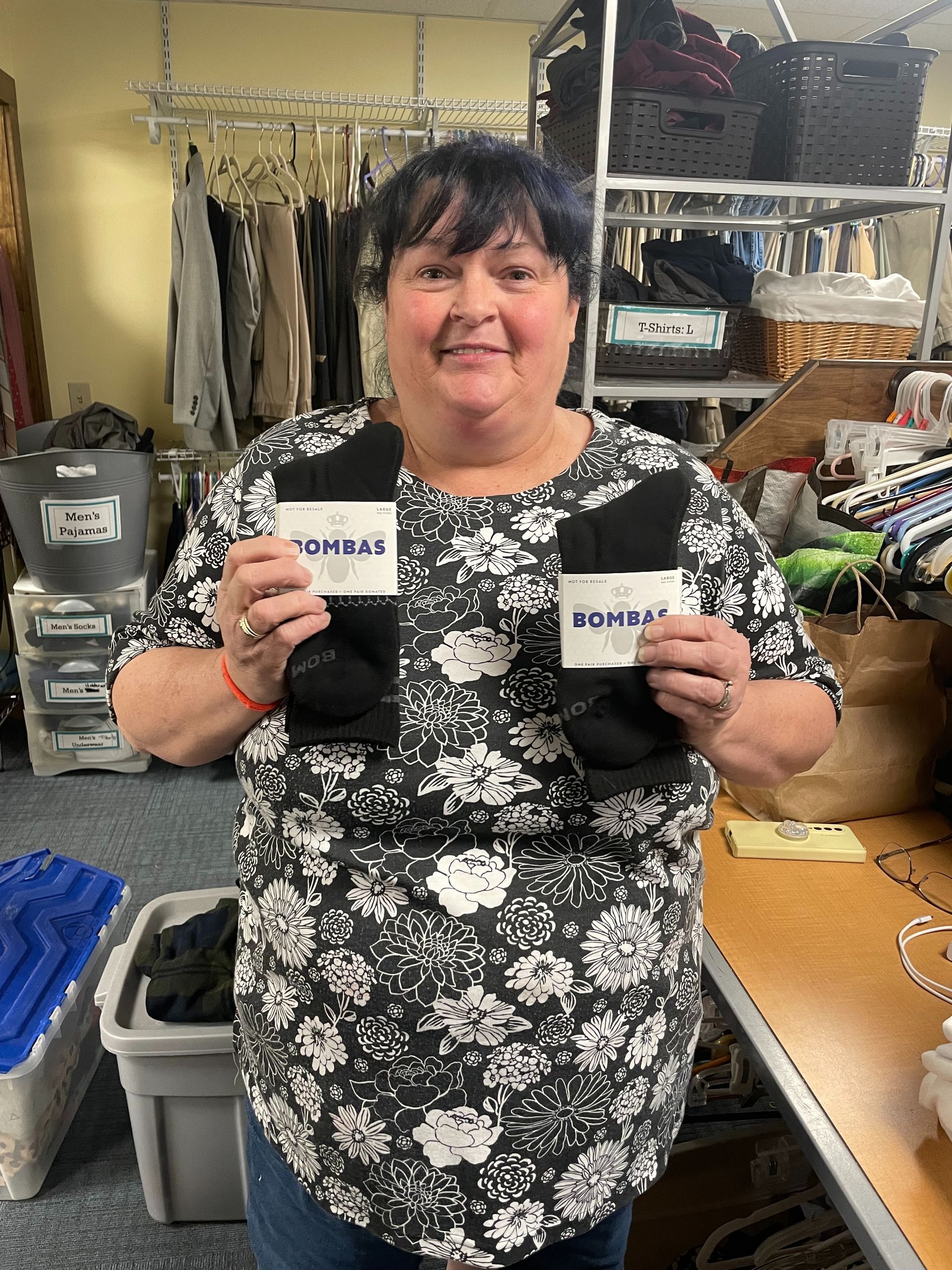 A person in a floral top smiles while holding up a pair of black socks in a room filled with clothing racks and storage.