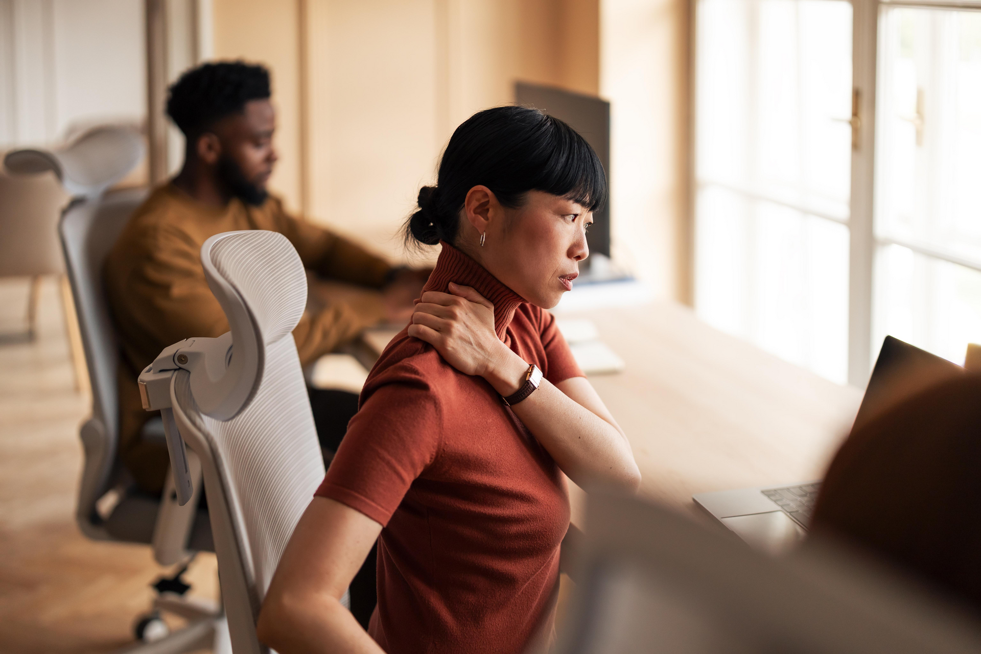 Person sitting at a desk with poor posture experiencing neck and lower back discomfort during computer work.