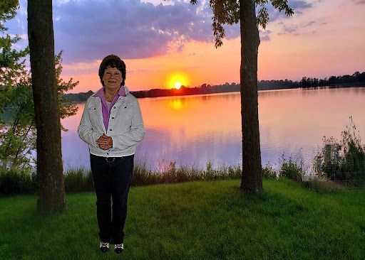 Person standing by a lake at sunset between trees, with colorful sky reflected in the water