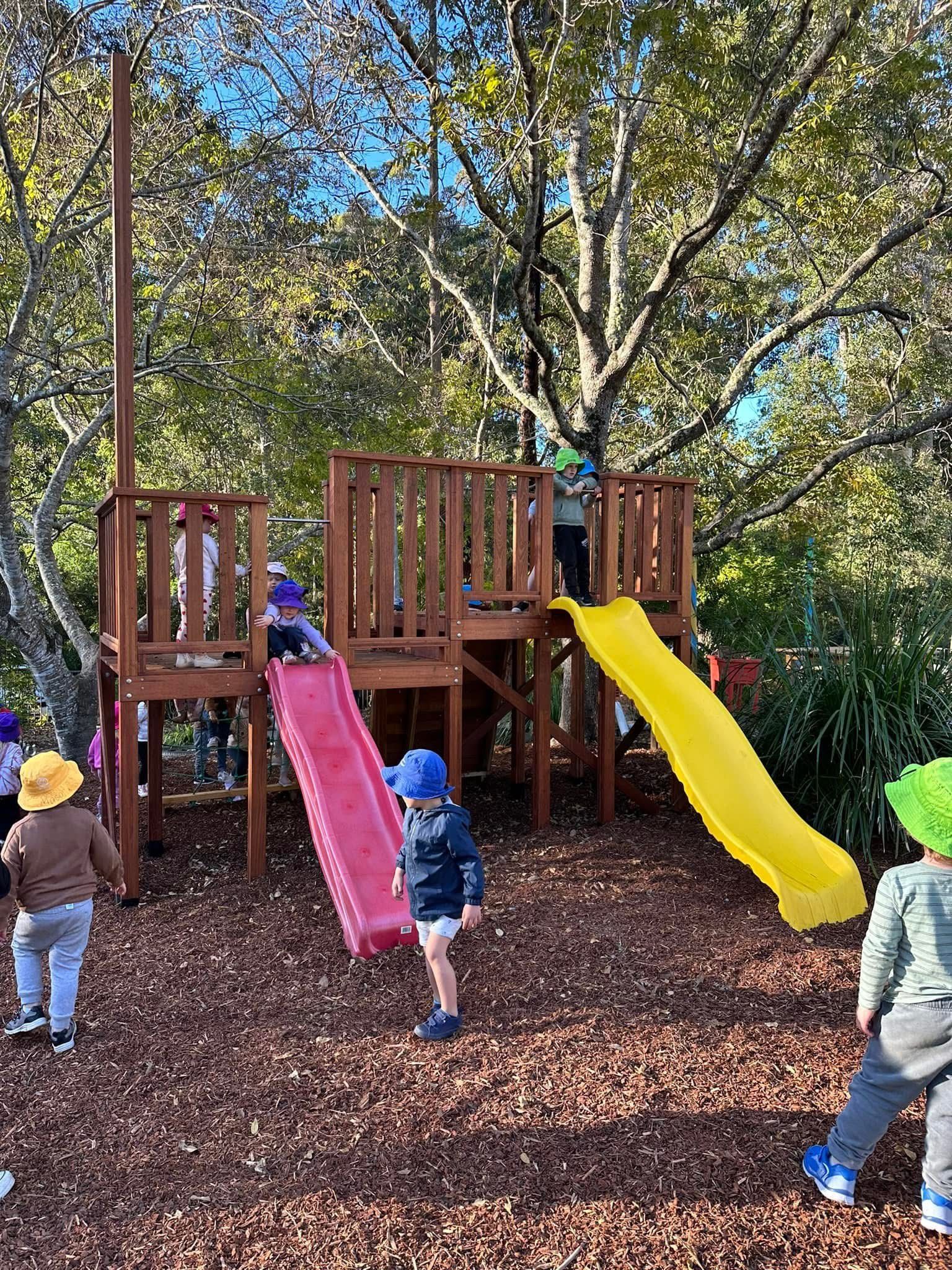 Children play on a wooden playground structure with a red slide and a yellow slide in an outdoor, wooded setting.