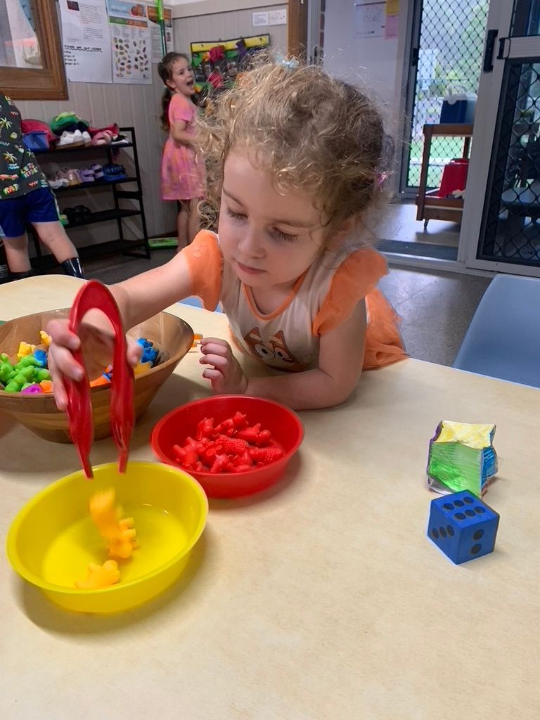 A child uses red tongs to pick up yellow animal figures from a bowl at a table with other bowls, a die, and a toy.