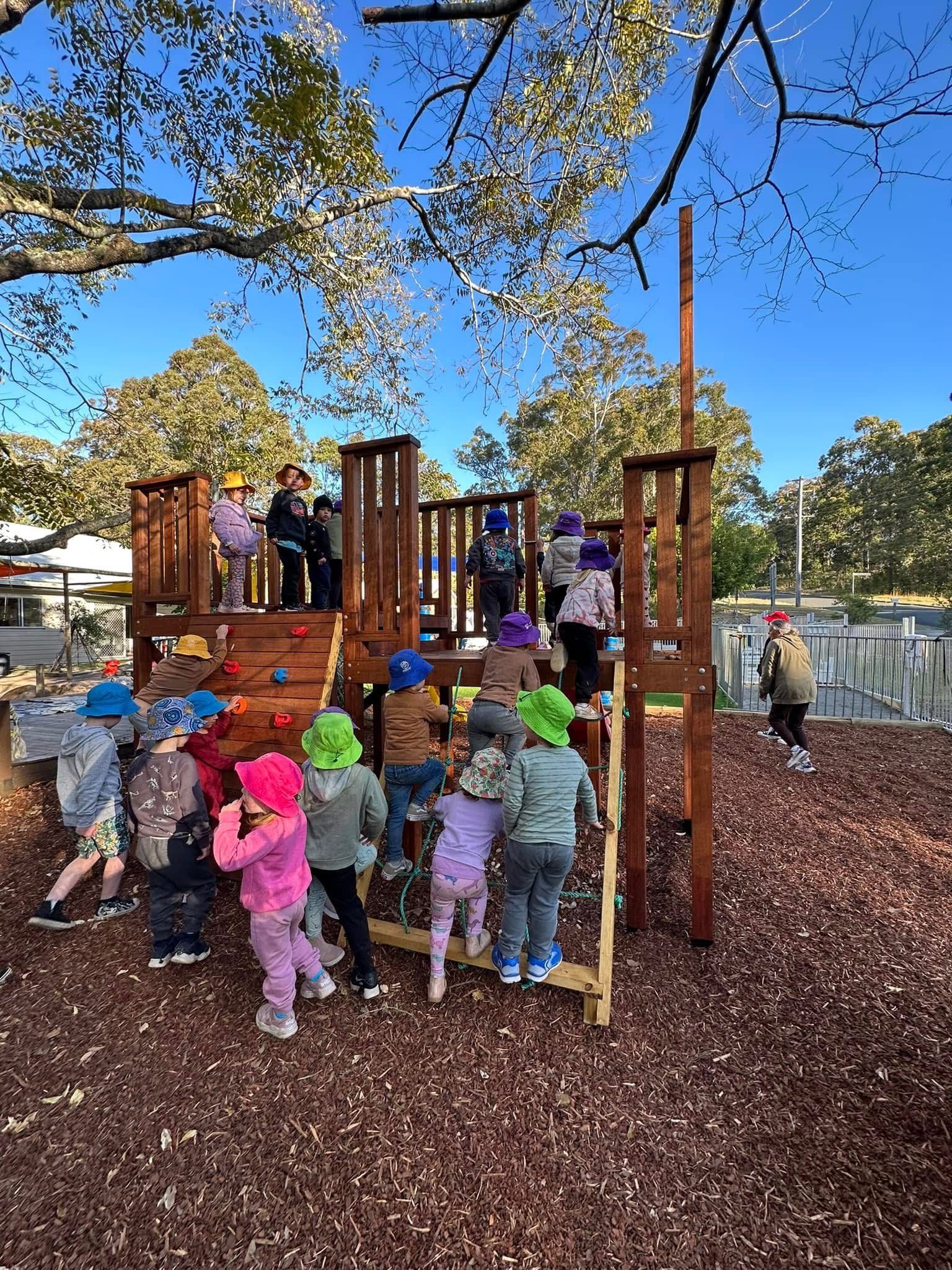 Children in colorful sun hats climb and play on a wooden playground structure on a sunny day with woodchip ground.