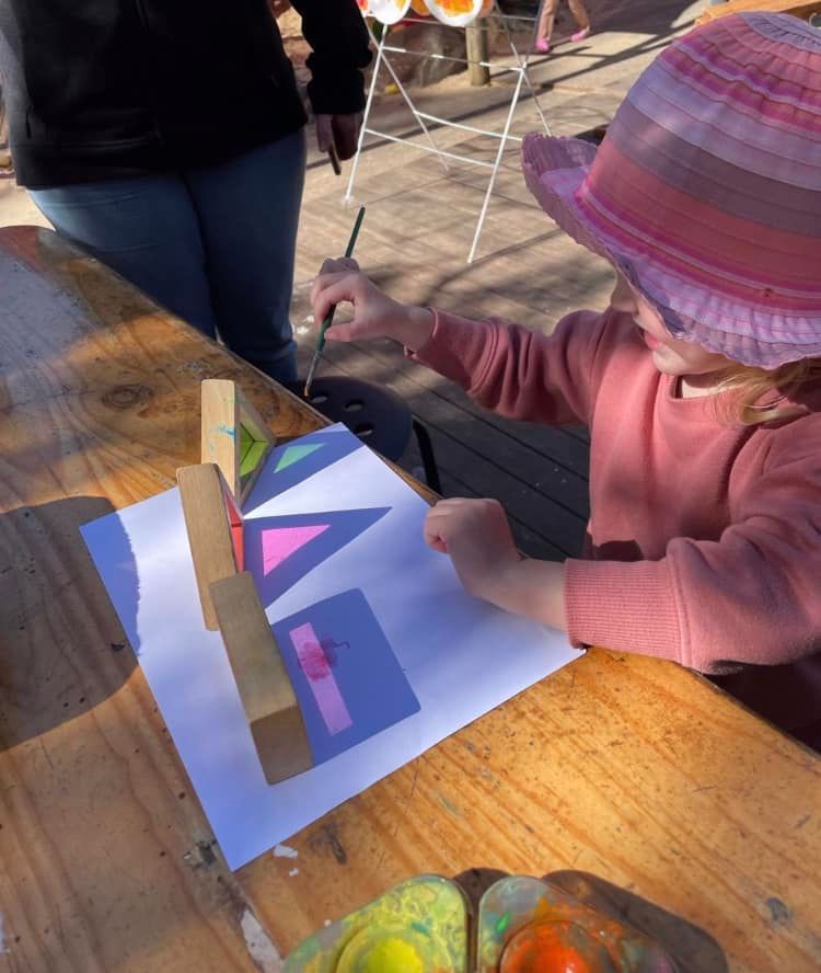 A child wearing a pink hat paints colorful geometric shapes on paper placed on a wooden table.