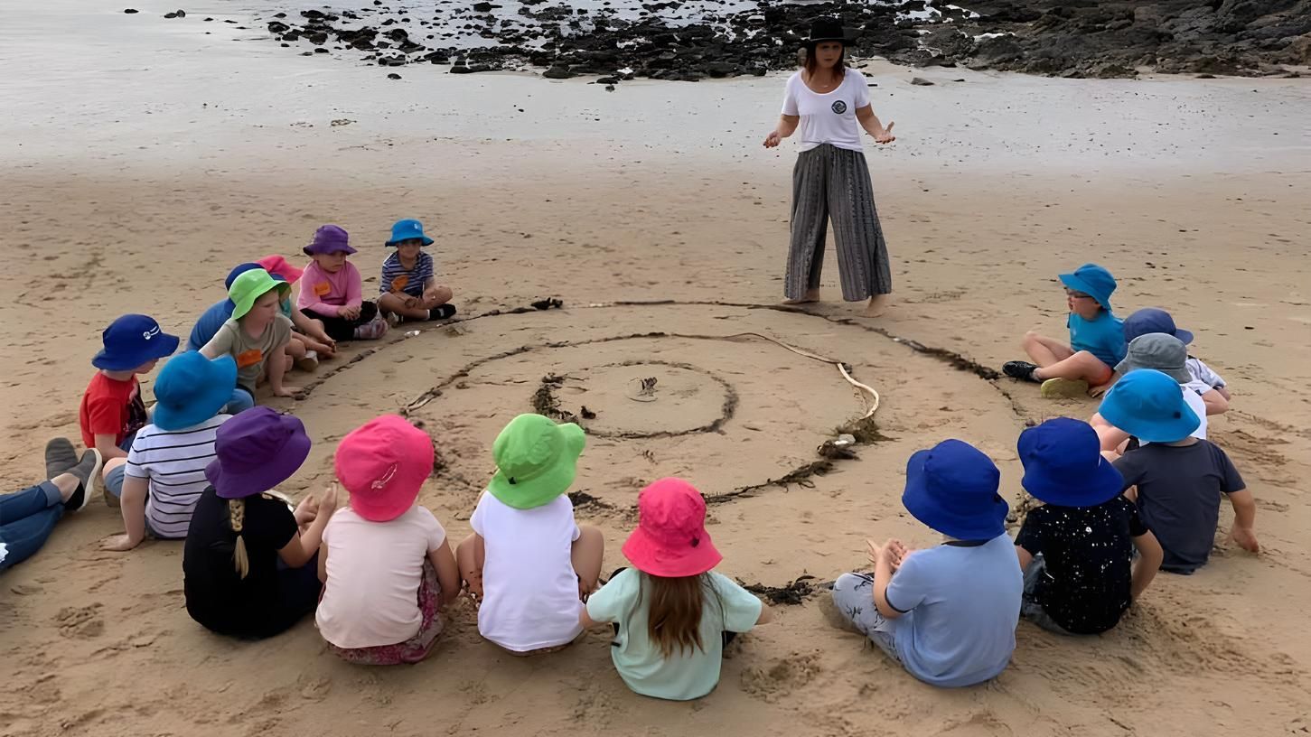 A group of children sits in a circle on the beach, surrounding markings drawn in the sand as an adult stands nearby.