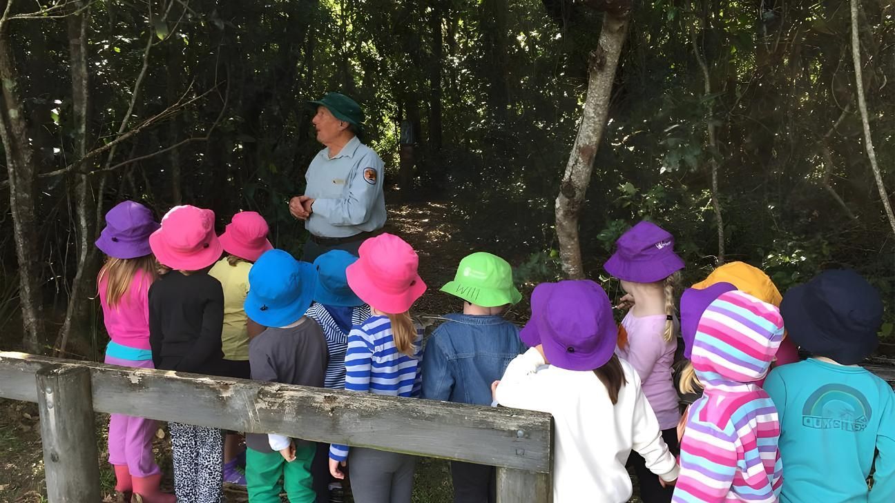 A park ranger talks to a group of children wearing colorful sun hats at the edge of a wooded area.