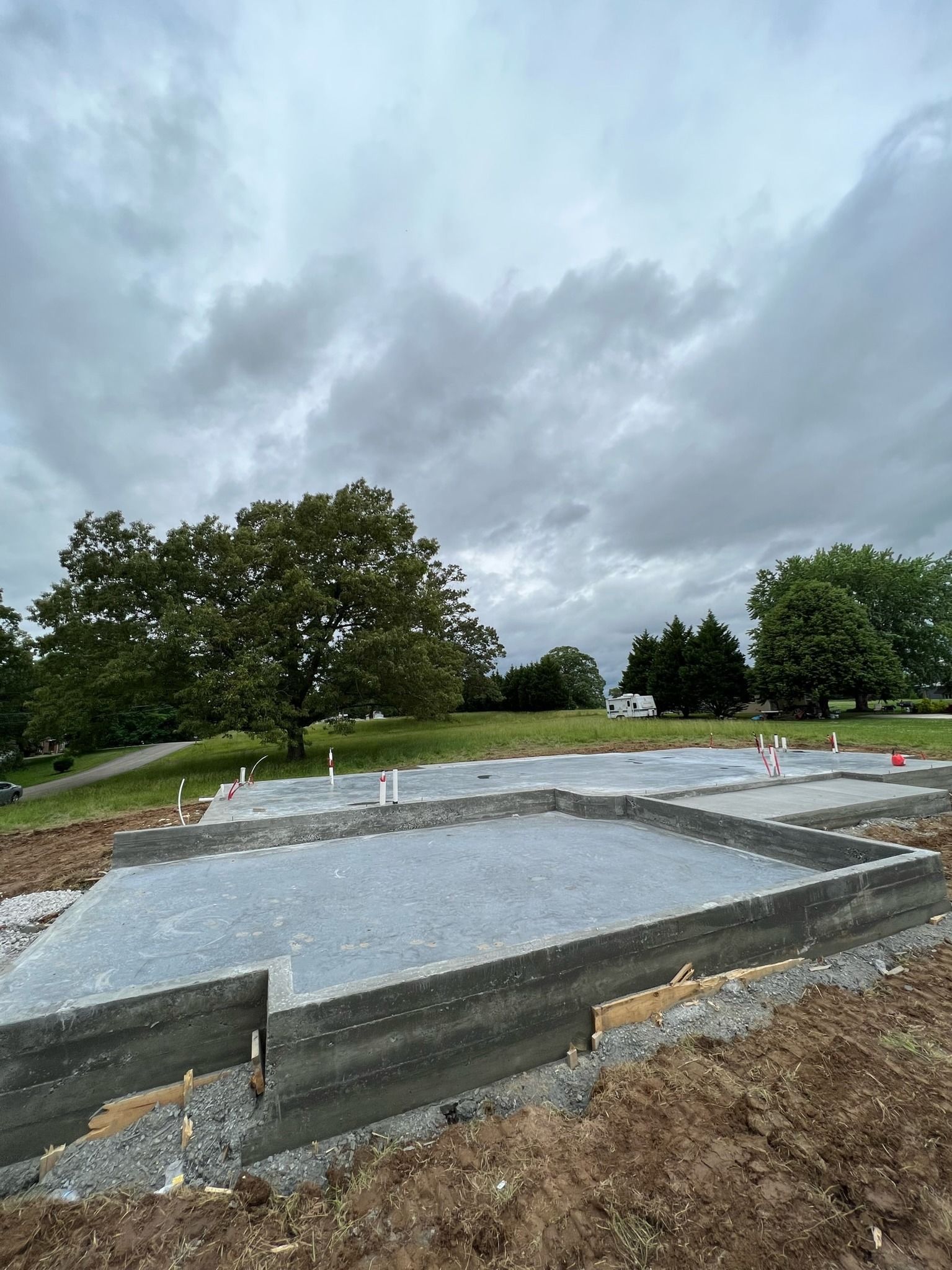 Construction site with a concrete foundation under an overcast sky; trees in the distance.