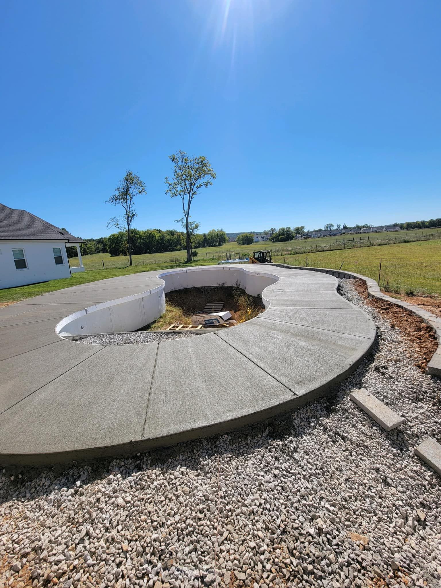 Concrete pool foundation under construction with gravel surround, green field, and blue sky.