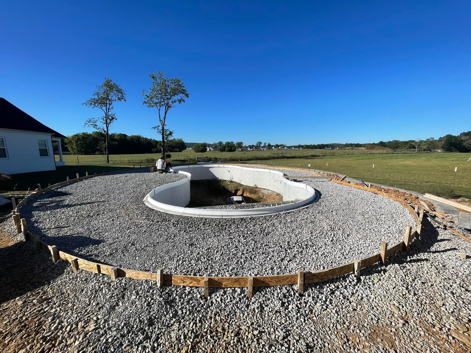 Construction site with a half-finished, kidney-shaped pool, surrounded by gravel, under a blue sky.