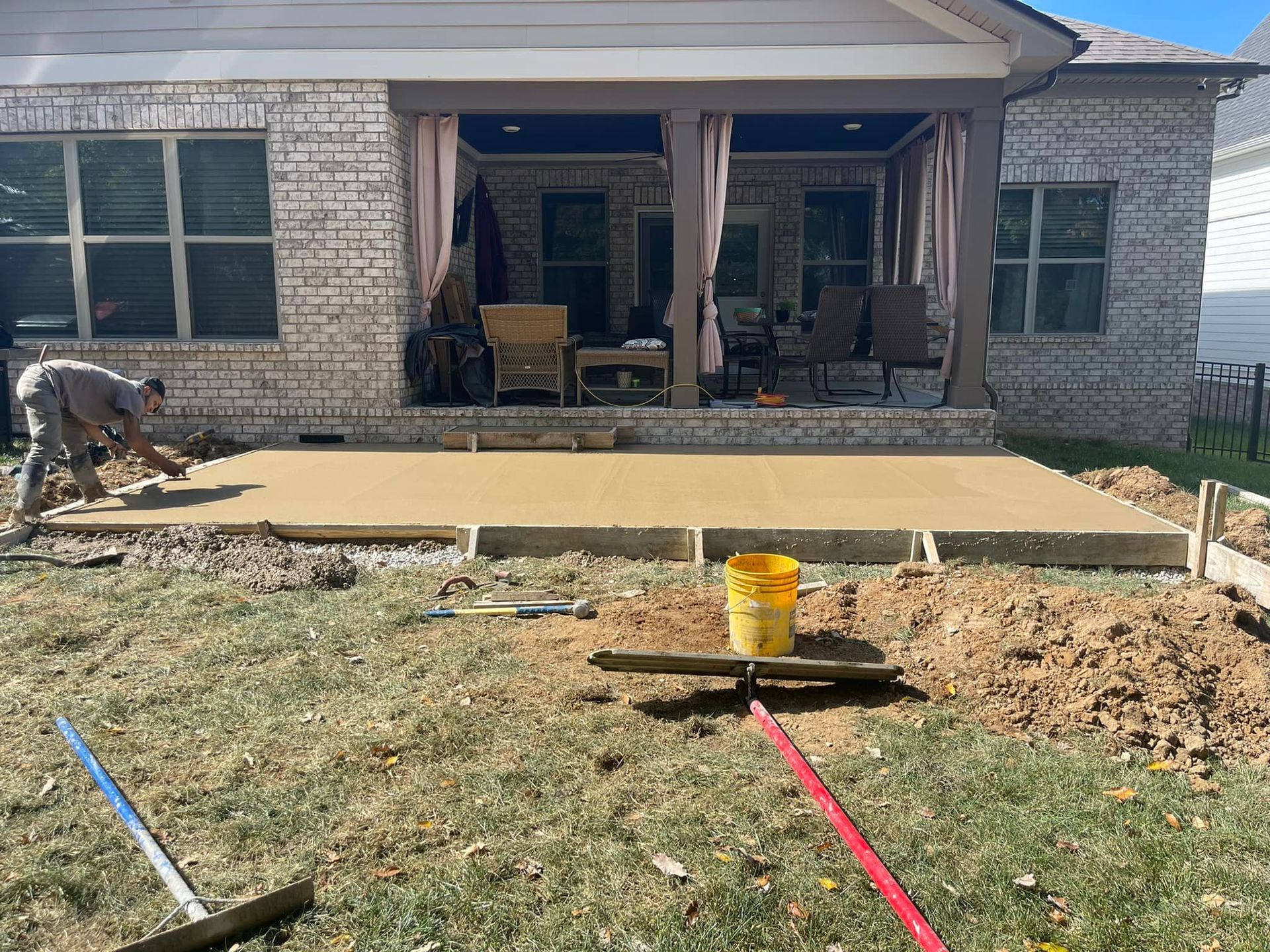 Construction worker pouring concrete for a patio in a backyard. The building is brick and has a covered porch.