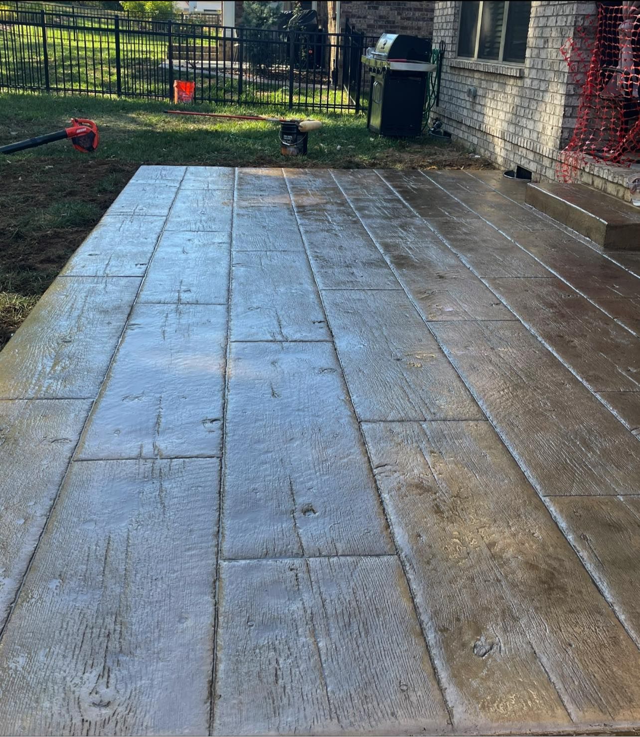 Stamped concrete patio with a wood-plank design. The gray surface is wet, and a leaf blower sits nearby.