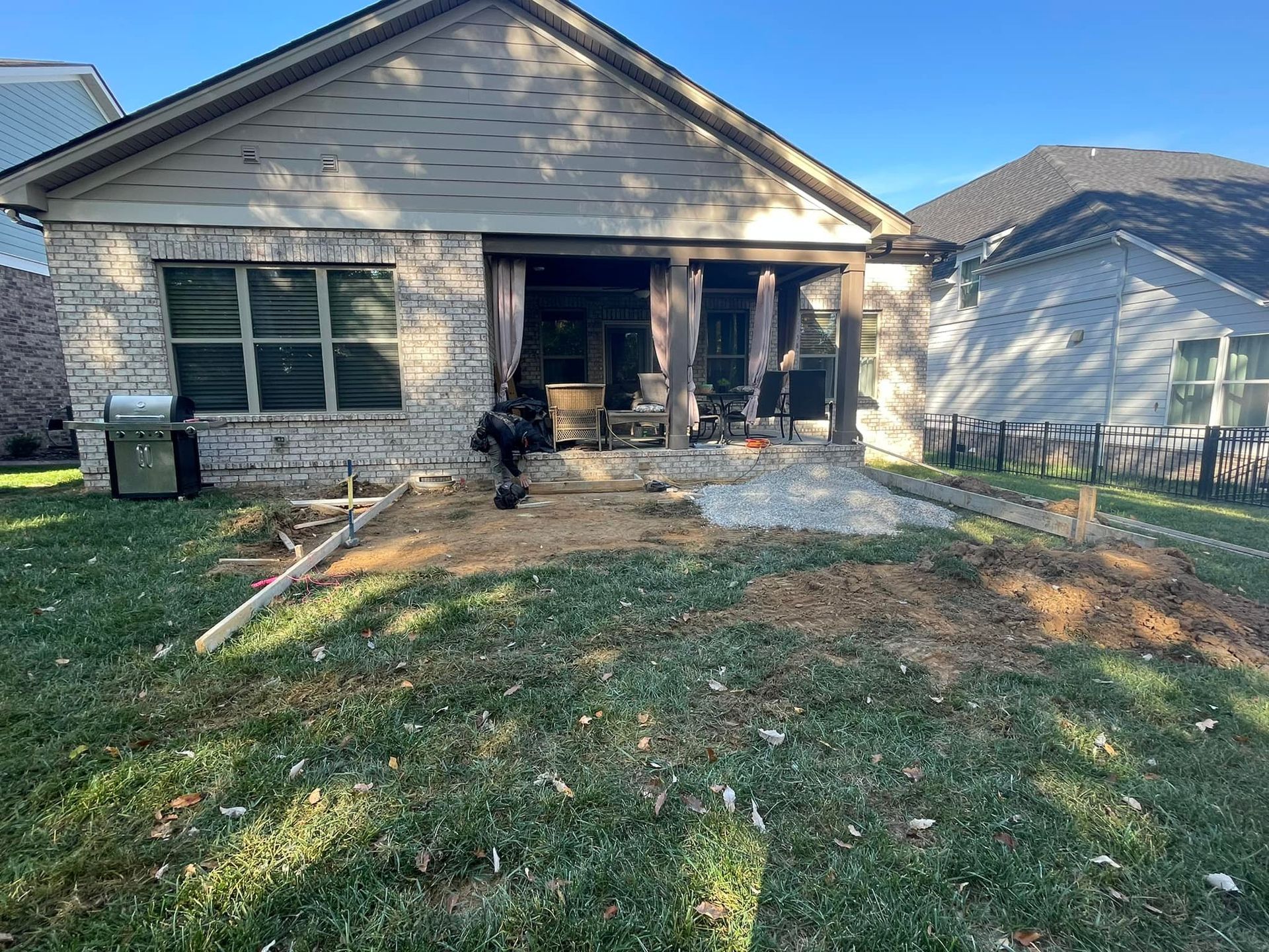Backyard with a person working on a patio. House has stone siding and a covered porch with curtains.