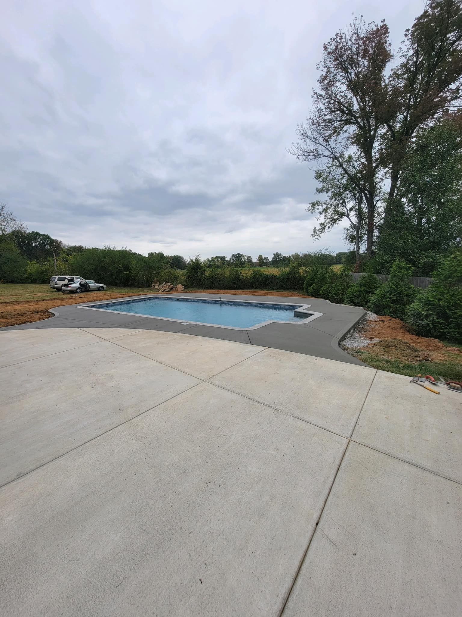 A concrete patio leads to a blue-tiled pool surrounded by dark concrete under an overcast sky.