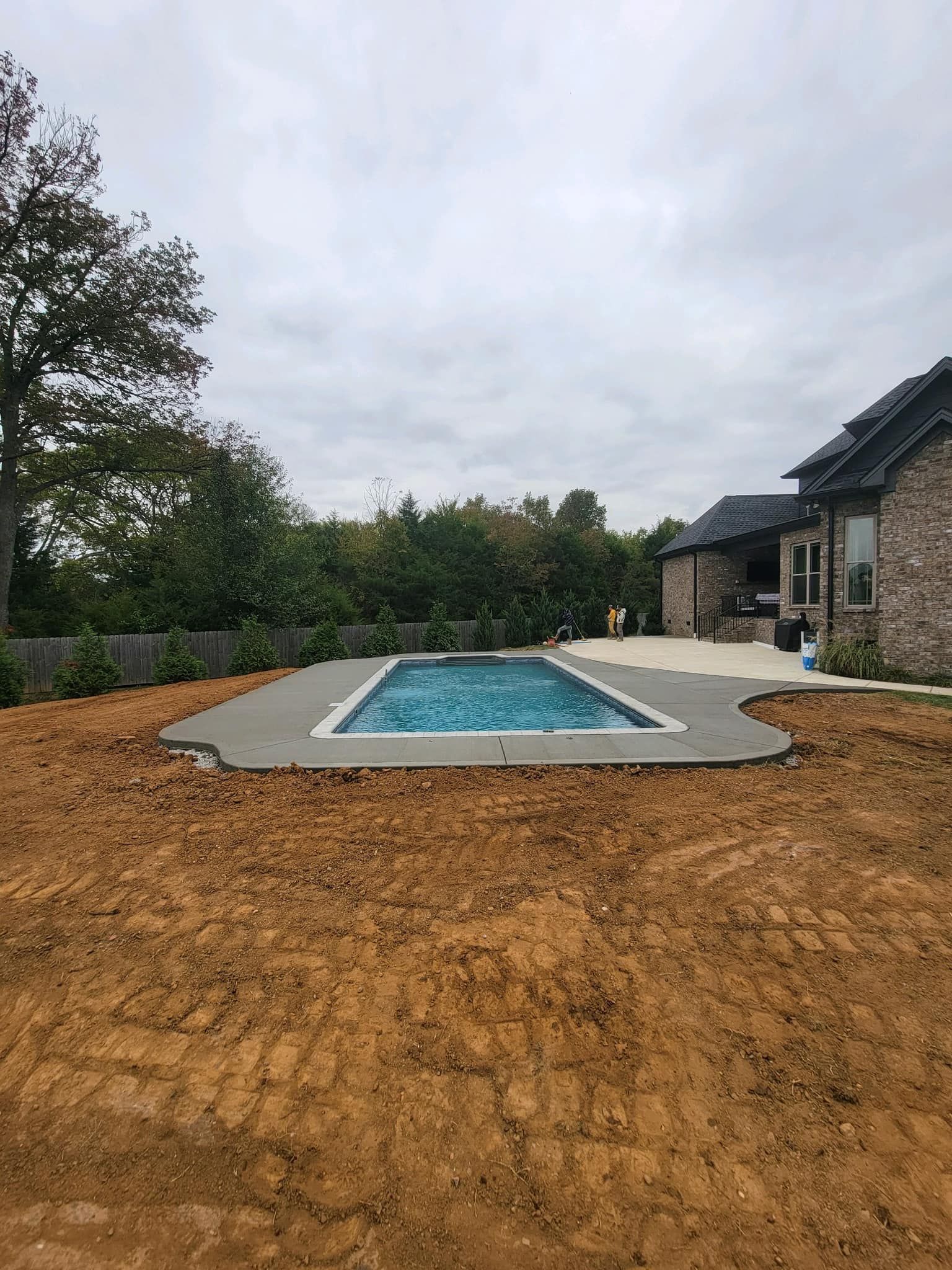 A rectangular pool with a gray concrete deck sits in a yard next to a brick house under a cloudy sky.