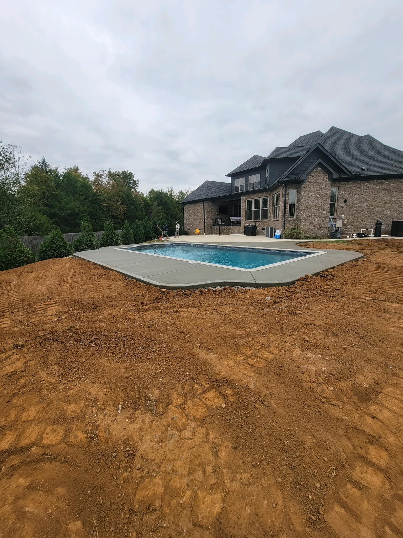 A partially-filled swimming pool surrounded by dirt, in front of a large house with a dark roof.