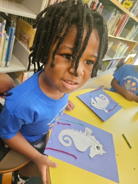 Child with dreadlocks smiles, holding a seahorse art project. They wear a blue shirt, seated at a yellow table.