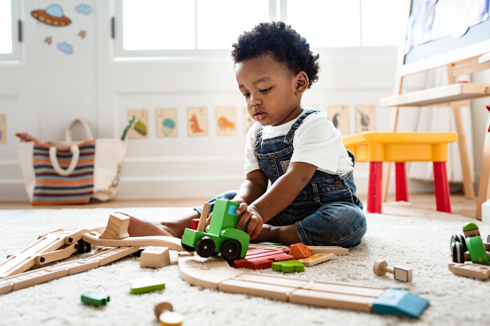 Young child in denim overalls playing with wooden train tracks and toy tractor on a carpeted floor.