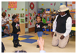Children in a classroom, one pointing, interacting with a person wearing a safari hat.