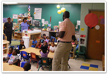 Man speaking to a group of children in a classroom. Two women stand behind them. Tables and chairs.