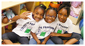 Three children in white shirts smile, sitting together. They are inside a room with shelves.