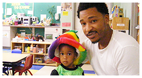 Man and child in a colorful hat in a classroom, smiling at the camera.