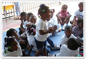 Children sitting in a circle, one standing, interacting outdoors.