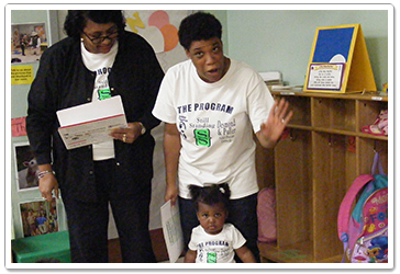 Two adults and a child in a classroom; one adult waves, the other reads a paper.