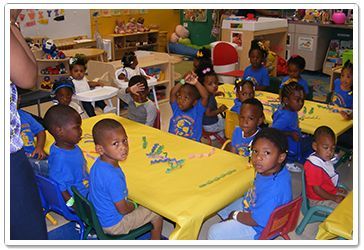 Children in blue shirts sit at tables with toys on them in a brightly lit classroom.