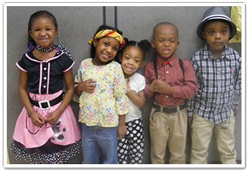 Five children in vintage-style outfits smiling. Standing against a wall.