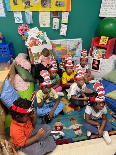 Children wearing Cat in the Hat hats sit on a rug in a classroom, reading.