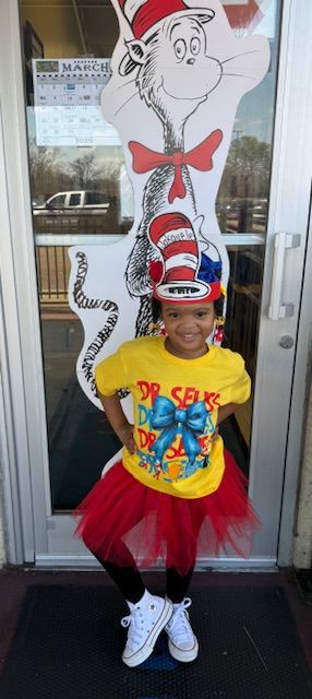 A girl in a Dr. Seuss costume poses in front of a window with a Cat in the Hat cutout.