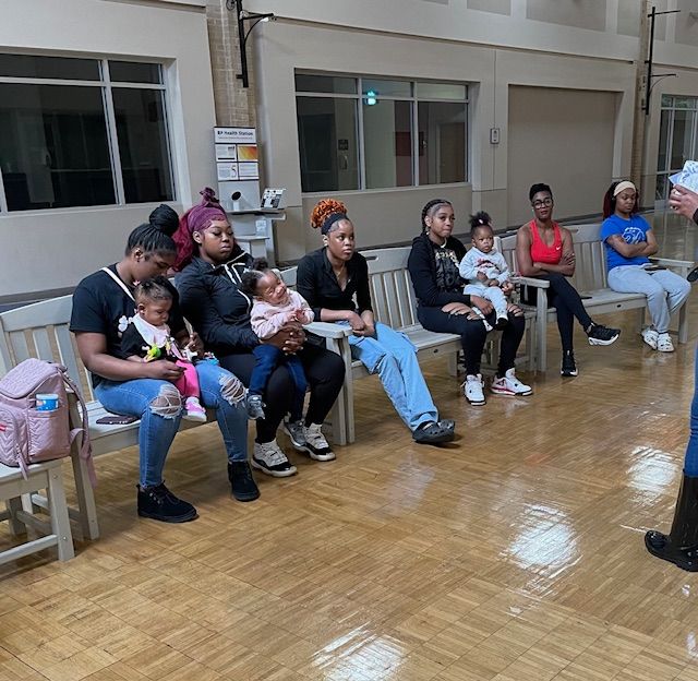 Group of women with babies sit on benches inside a building, possibly a waiting area.