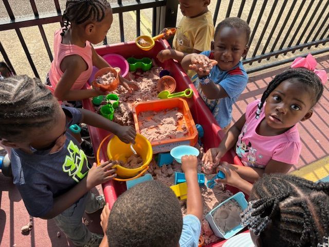 Children in blue shirts at a table playing with blocks in a bright classroom.