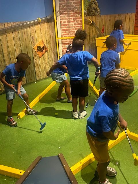 Children in blue shirts playing mini golf on a green course with yellow borders.