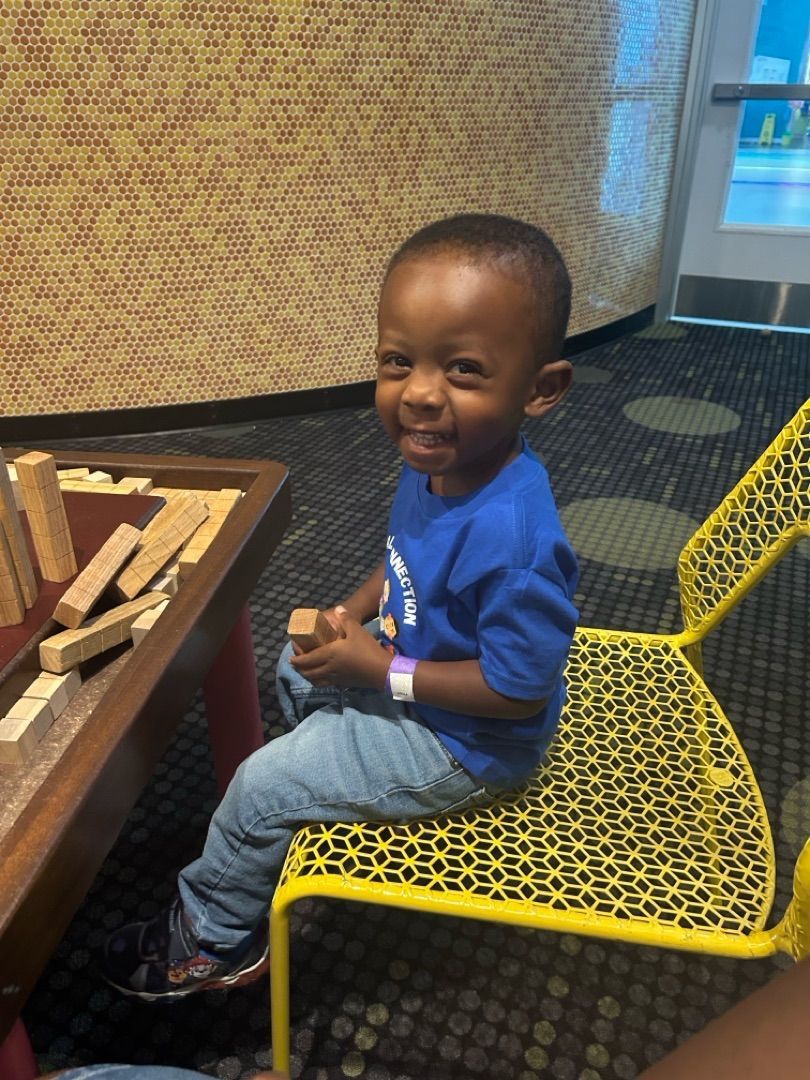 Children seated on a rug in a classroom, smiling at the camera. Some are waving.