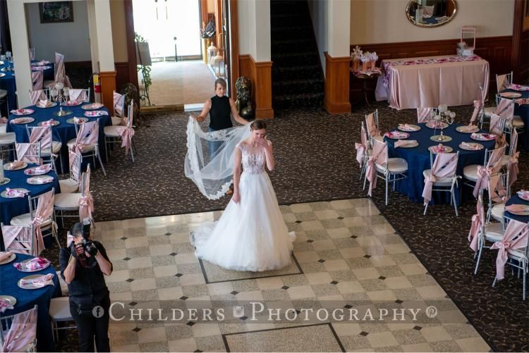 Bride in wedding dress walks on checkered floor, trailed by attendant, in a decorated event space with blue tables.
