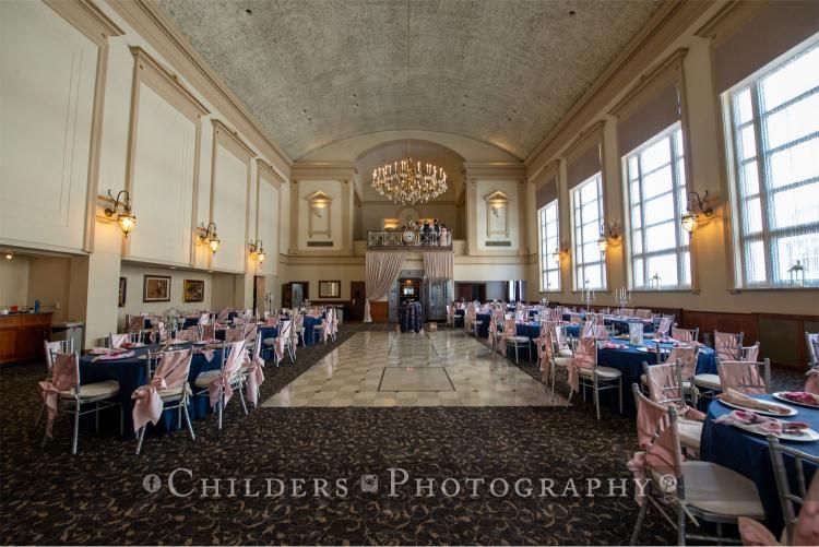 Formal event hall with round tables, navy tablecloths, pink chair sashes, and a central dance floor.