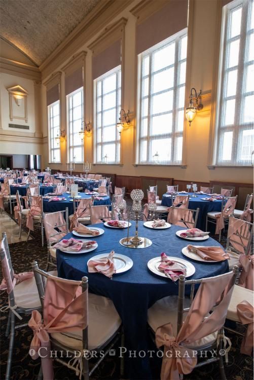 Formal event room with round tables, blue tablecloths, pink accents, and large windows.