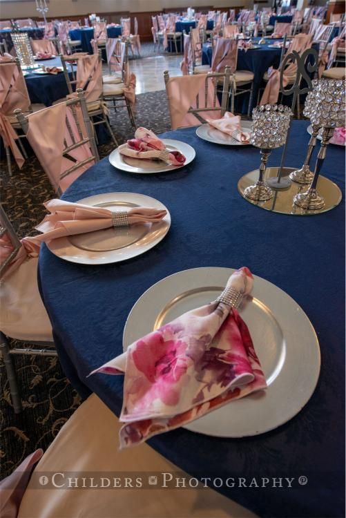 Navy blue table set with silver charger plates, pink floral napkins, and pink chair covers in banquet hall.
