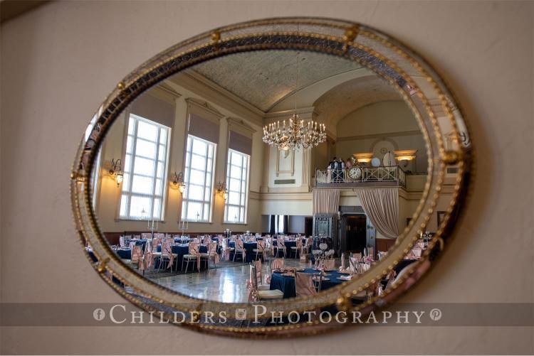 Oval mirror reflecting a ballroom with tables set for an event; windows and chandelier are visible.