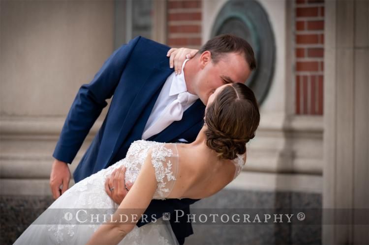 Groom in blue suit kisses bride in white dress; they are outside a brick building, a romantic embrace.