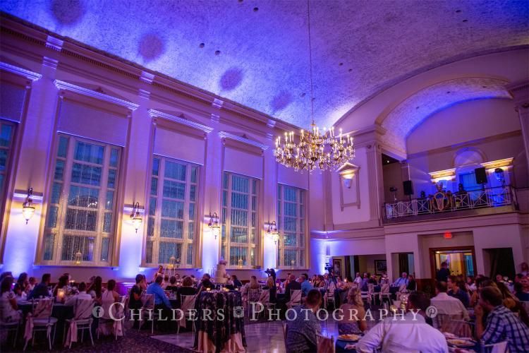 Event hall with blue lighting, people at tables, band on balcony, ornate chandelier.