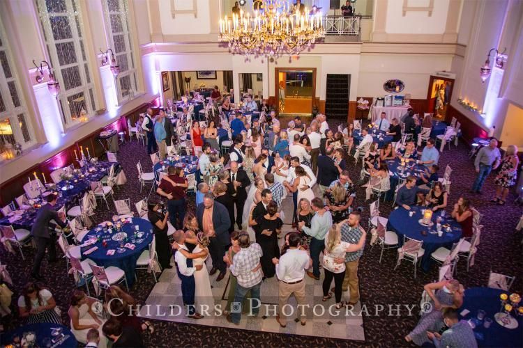 Wedding reception in a large hall with guests mingling around tables, dancing on a floor, and under a chandelier.