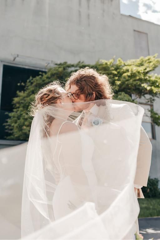 Newlyweds kissing, veil blowing in wind. Bride in white dress, groom in suit, outdoors.