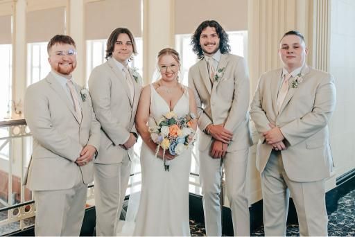 Bride and groomsmen in a tan suits, posing for wedding photos. Inside building with a staircase and windows.