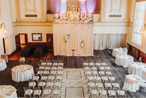 Ceremony hall set up with chairs, tables, a draped backdrop, and chandelier.