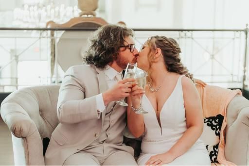 Bride and groom kiss while toasting with champagne; they sit on a cream-colored couch in a luxurious setting.