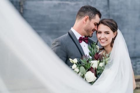 Groom and bride smiling, holding bouquet, outdoors. Bride in veil. Groom in gray suit, burgundy bow tie.