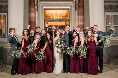 Wedding party celebrating outside a building; couple kissing, bridesmaids in burgundy dresses, men in suits.