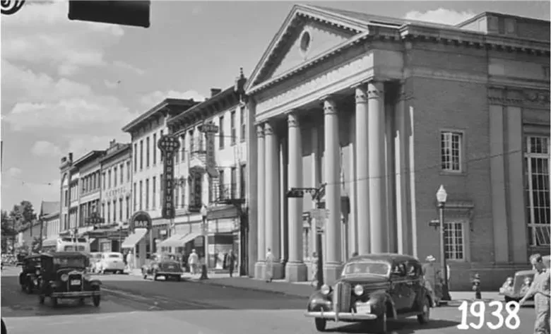 A black and white street scene from 1938, with cars and pedestrians, and a large building with columns on the right.