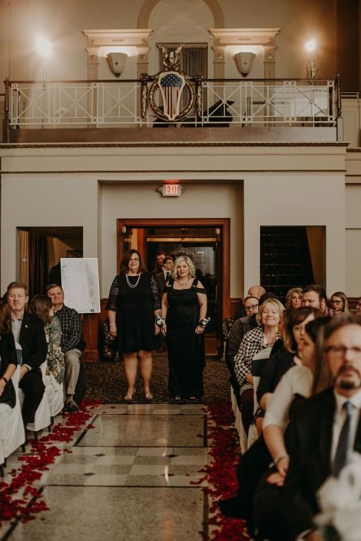 Two women walk down aisle lined with rose petals, towards wedding ceremony. Guests seated on both sides.